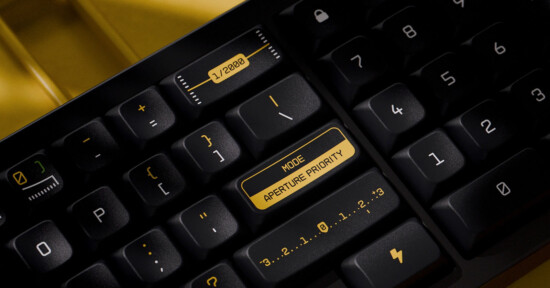 Close-up of a mechanical keyboard with black keys and yellow text. One key reads "MODE APERTURE PRIORITY," while others show various icons and numbers, including a camera aperture symbol and a lightning bolt.