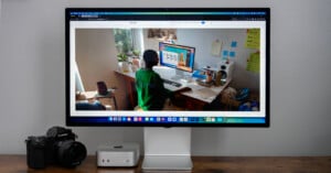 A computer monitor displays an image of a person working at a desk in a room filled with plants and various items. Below the monitor, a camera and a small computer device are placed on a wooden surface.