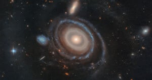 A luminous spiral galaxy with distinct rings glows against a backdrop of countless stars and other galaxies. Its bright core is surrounded by swirling blue and white patterns, creating a mesmerizing cosmic display.