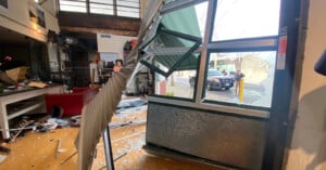 Interior scene of a damaged room with debris and shattered glass scattered on the floor. A damaged window frame is leaning inward. Outside, a police car is parked on the street. A person stands inside, surveying the damage.