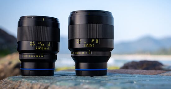 Two black camera lenses with yellow marking details stand on a stone surface. The background features a soft-focus landscape with mountains and a clear sky.