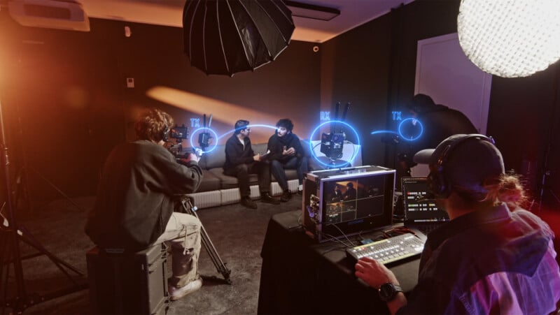 A video production setup with two people sitting on a couch being filmed. A camera operator is capturing the scene, and a person in front of a control panel monitors the recording. Professional lighting and equipment surround them in the studio.