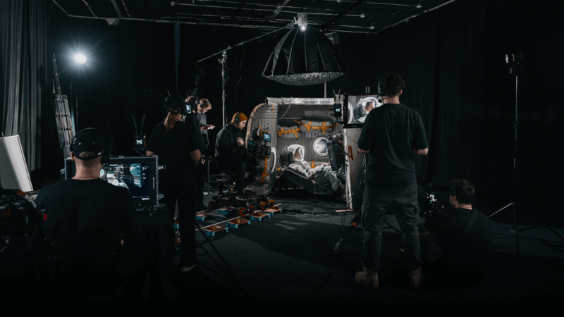 Film crew capturing a scene with an actor in a space suit inside a spacecraft set. There are cameras, lighting equipment, and crew members adjusting settings in a dark studio environment.