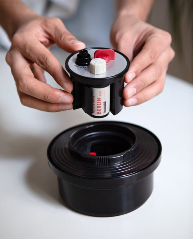 Hands assembling a black film developing tank with a roll of film labeled "BERLIN 400" inside. The tank has a removable lid with two knobs, one black and one red. The setup is on a light surface.