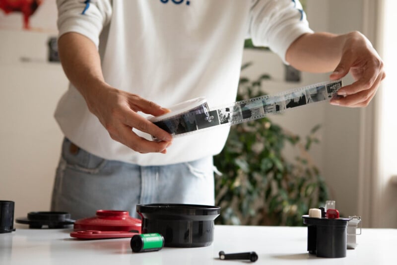 Person handles a strip of photographic film in a room, with film canisters and developing equipment on the table. They're wearing a white shirt and jeans. A plant and blurred wall art are in the background.