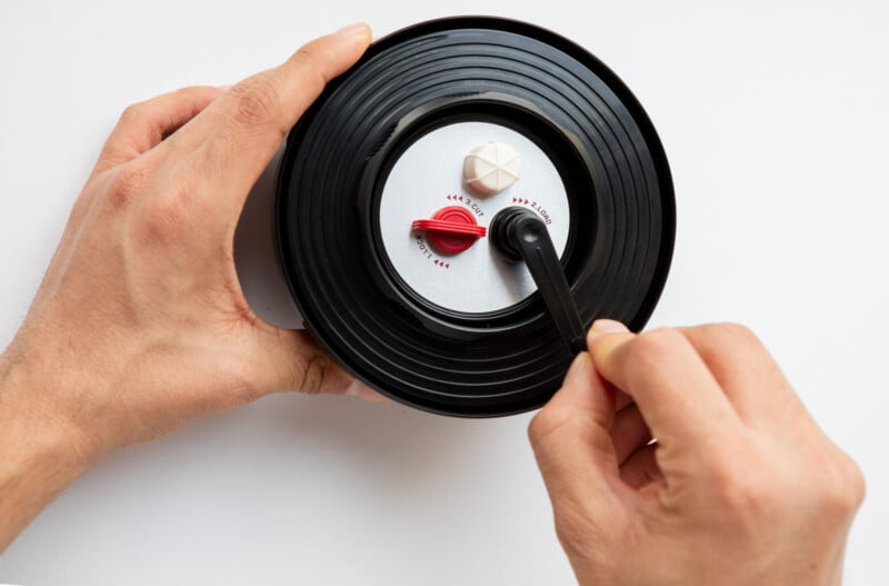 Two hands are holding a round black gadget with a knob and a rotating handle. The device resembles a mini record player. One hand is turning the handle, while the other stabilizes it, against a plain white background.