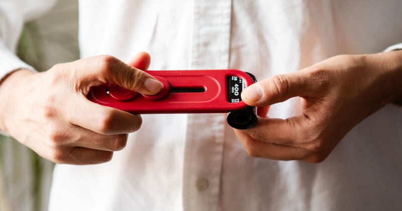 A person in a white shirt is holding a red handheld device with a small screen displaying numbers. The device has a lens attachment on one end, suggesting it may be a camera or similar gadget.