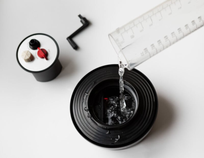 A clear liquid is being poured from a graduated cylinder into a black film developing tank, which has its lid off. Another tank lid and a small handle are visible in the background on a white surface.