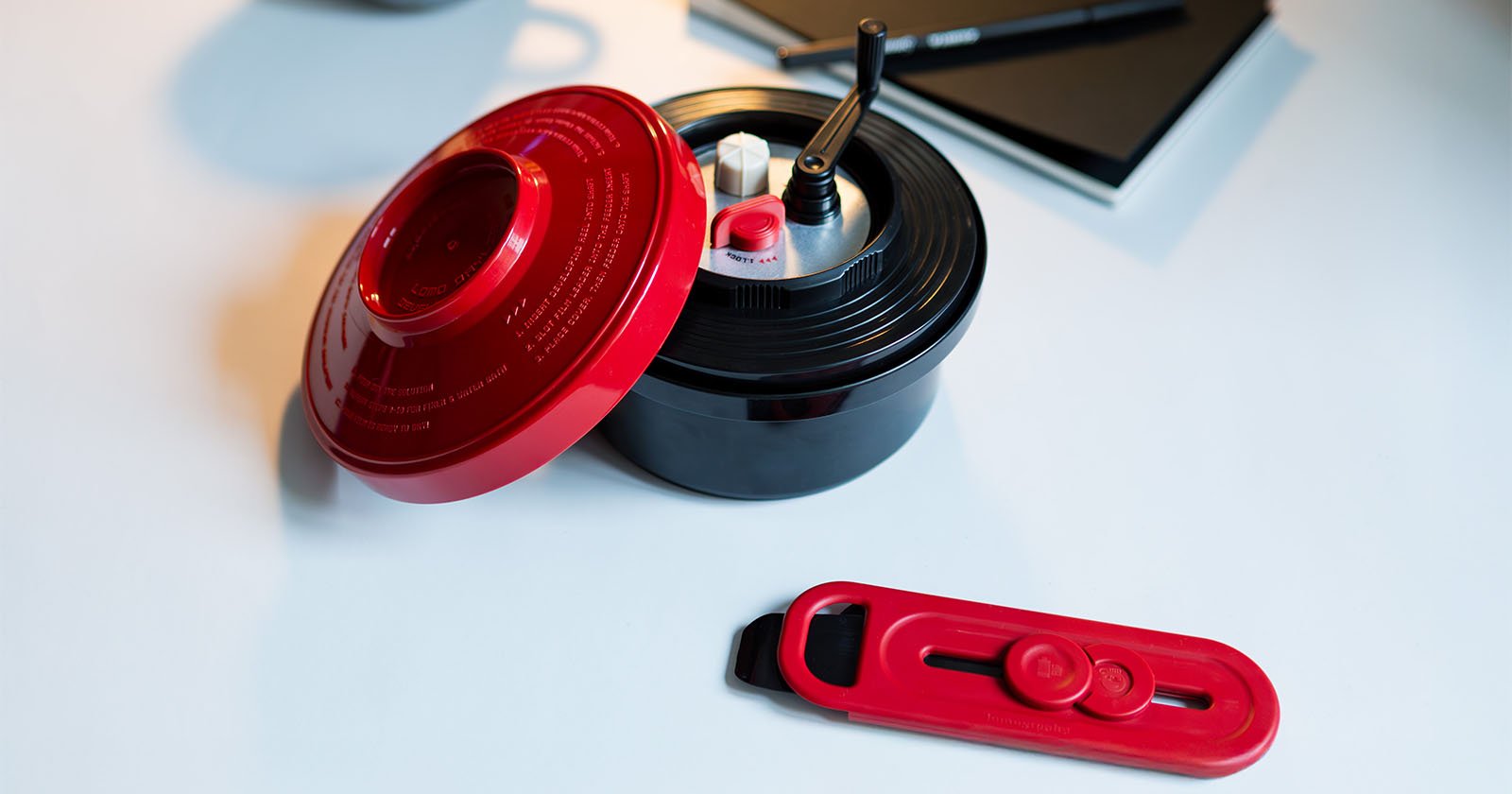 A red and black kitchen gadget with a lid and crank handle is on a light surface. A matching red attachment lies nearby. A notebook and pen are in the background.