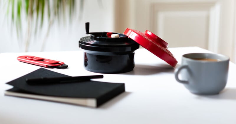 A darkroom developing tank with a red lid sits open on a white table, alongside a red box cutter, a black notebook with a pen, and a gray mug filled with coffee. A blurred green plant is in the background.