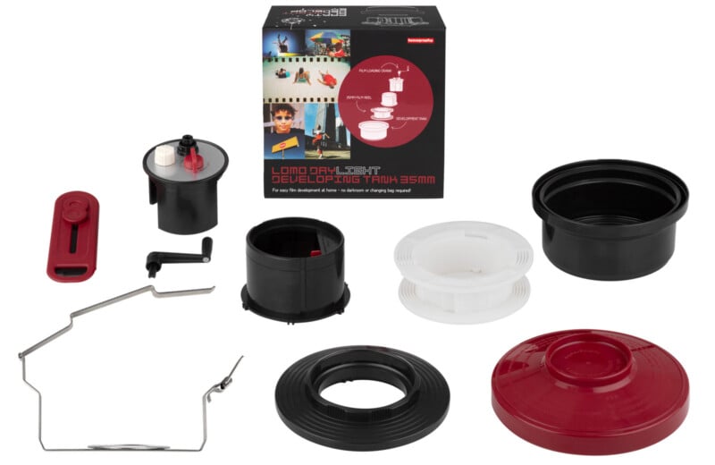 A Lomo daylight developing tank kit for 35mm film is displayed. The set includes a black tank, red lid, reels, a film squeegee, and a metal film opener. The product box shows images of film development.