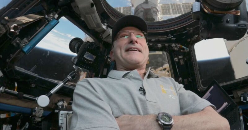 A person wearing glasses, a cap, and a light gray shirt stands inside a spacecraft cockpit with visible controls, monitors, and a window showing the sky and parts of the space station outside. The person is looking upward and smiling.