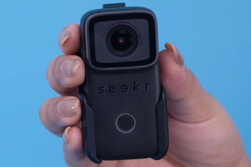 A hand holding a black rectangular camera with a lens at the top. The word "seekr" is engraved below the lens. The background is a solid light blue.