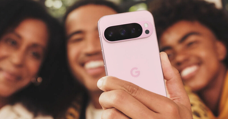A person holds a pink Google Pixel phone with the camera facing outward, capturing a selfie. Two people smile in the background. The phone's "G" logo is visible on the back.