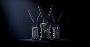 Three black wireless transmitter units with multiple antennas are standing on a dark surface against a black background. The central unit displays a digital screen with channel information.