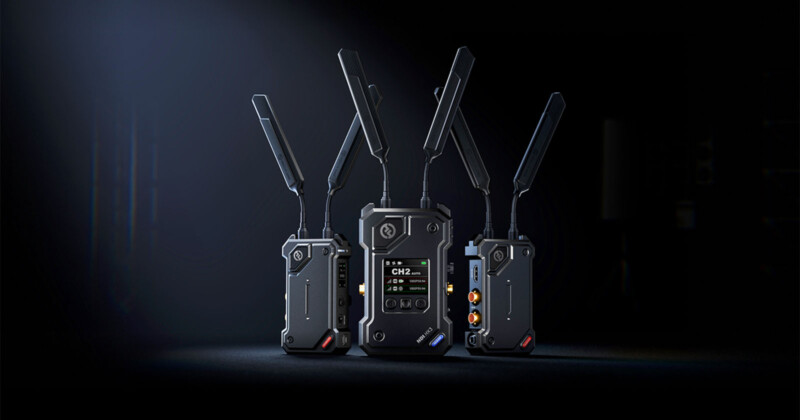 Three black wireless transmitter units with multiple antennas are standing on a dark surface against a black background. The central unit displays a digital screen with channel information.