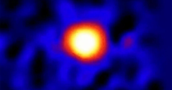 A blurry infrared image of a celestial object, with a bright yellow and orange center surrounded by a diffuse blue glow, possibly representing a distant star or planet.