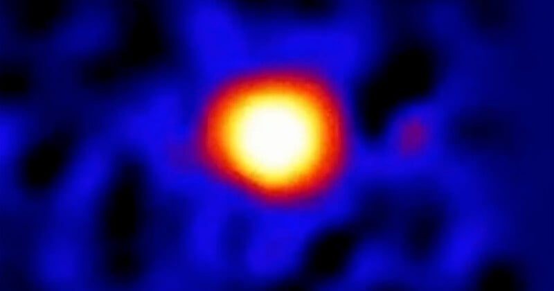 A blurry infrared image of a celestial object, with a bright yellow and orange center surrounded by a diffuse blue glow, possibly representing a distant star or planet.