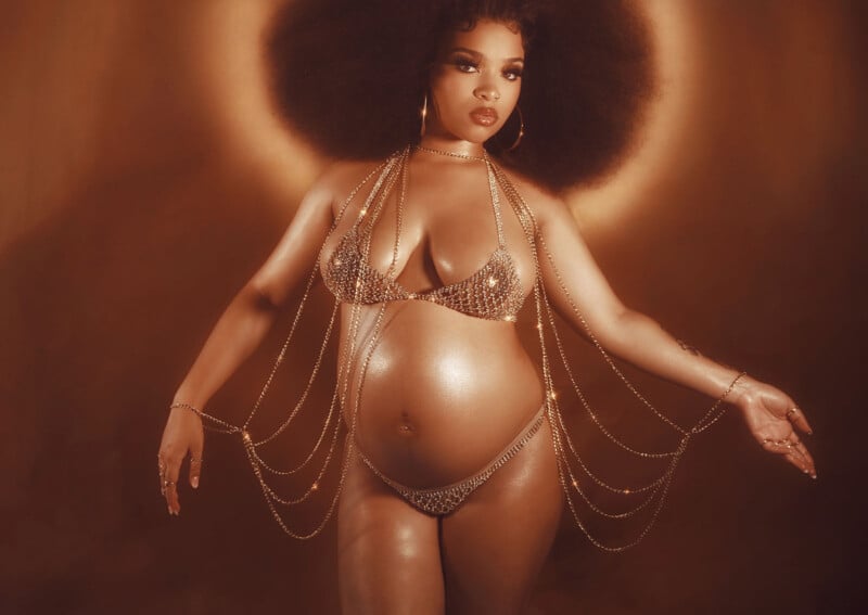 A pregnant woman stands confidently, wearing a sparkling bikini adorned with gold chains. Her hair is voluminous and styled in an afro. The background is a warm, golden hue, adding a radiant effect.