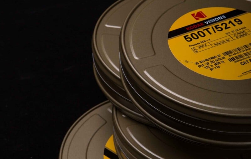 Close-up of stacked metal film canisters with yellow Kodak Vision3 labels. The labels display "500T/5219" and other technical details. The background is black, highlighting the canisters.