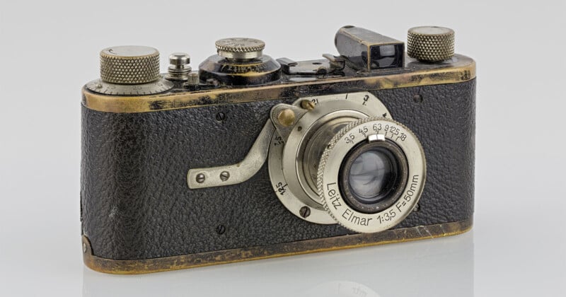 Vintage rangefinder camera with a well-worn brass and black body, featuring a prominent Leitz Elmar 1:3.5 f=50mm lens. The camera shows signs of age, including scratches and patina, set against a plain backdrop.