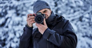 A person in a winter coat and gray beanie is holding a Leica camera up to their face, focusing on taking a photo. The background is snowy, providing a cold and wintry atmosphere.