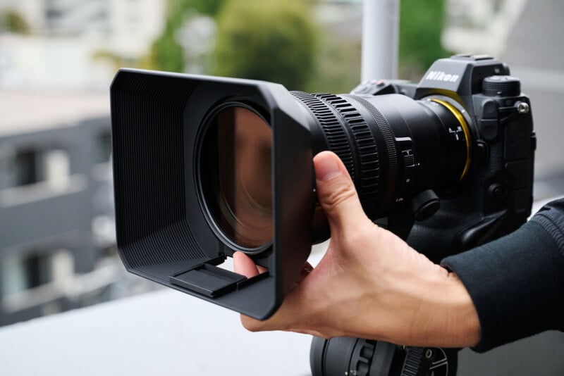 A person holds a DSLR camera with a large telephoto lens, featuring a square lens hood. The camera is pointed outdoors, showing part of a blurred cityscape background.