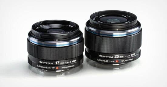 Two black camera lenses, labeled OM SYSTEM, are displayed side by side. The lens on the left is a 17mm f/1.8 and the one on the right is a 25mm f/1.8. Both lenses feature "SPLASH PROOF" labeling and have a sleek design.