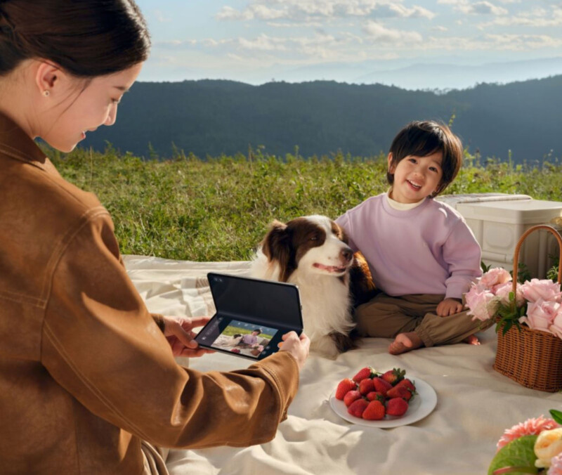 A woman takes a photo with a foldable phone of a smiling child sitting on a picnic blanket with a dog. There are strawberries and flowers on the blanket. They are outdoors with hills and a cloudy sky in the background.