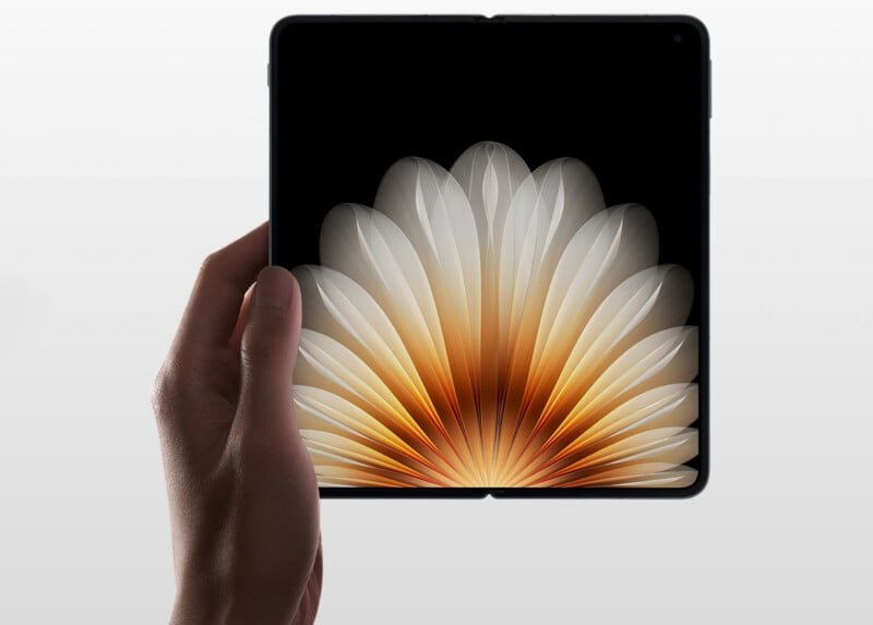A hand holding a foldable smartphone with its screen unfolded. The display shows an artistic image resembling a blooming flower in shades of white and orange. The background is a plain, light grey.