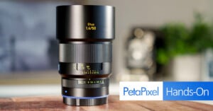 A Zeiss Otus 1.4/50 camera lens on a wooden surface. The lens is in focus with visible aperture and focus markings. The background is blurred, and there's a "PetaPixel Hands-On" graphic at the bottom right.