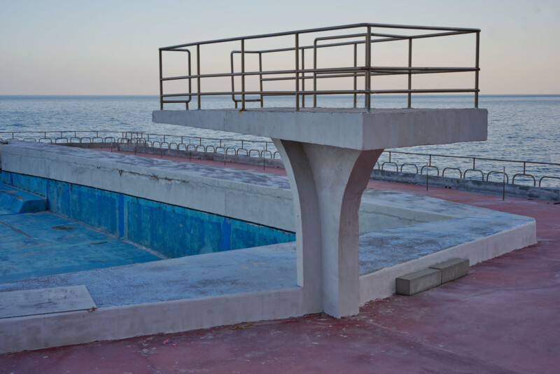 A concrete diving platform stands over an empty swimming pool with weathered blue paint. The ocean is visible in the background under a clear sky. The surrounding deck is reddish-brown, with metal railings along the pool's edge.
