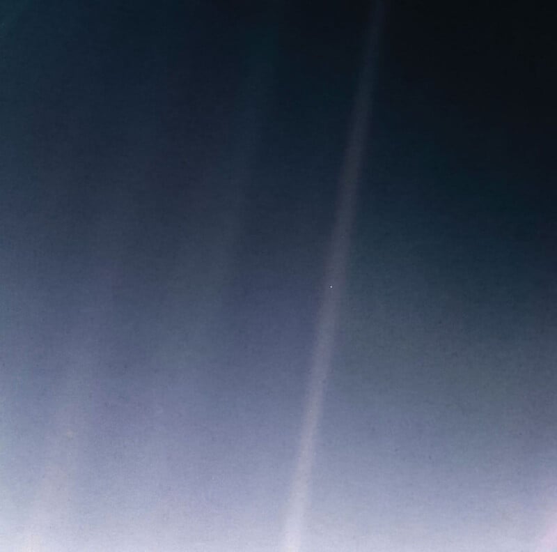 A photograph of space with a small pale blue point near the center, set against a dark, gradient background. Vertical light streaks are faintly visible, suggesting sunlight or camera glare.