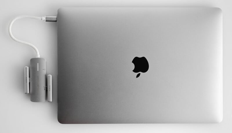 A silver laptop with an apple logo is connected to a gray USB-C hub adapter. The adapter has multiple ports and is plugged into the laptop on a light gray background.