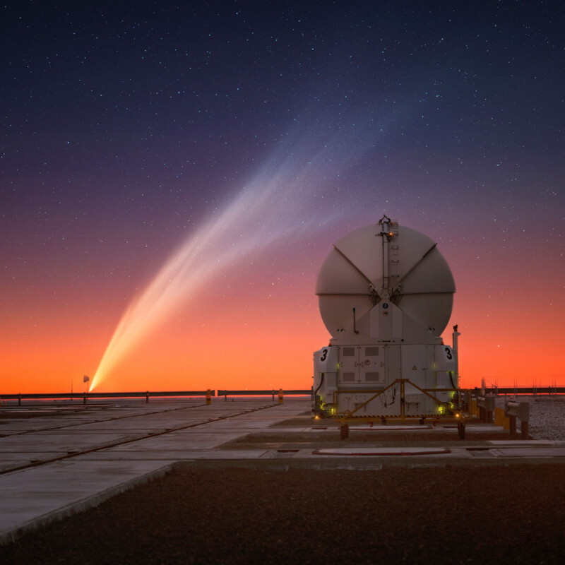 A large observatory telescope is positioned on a flat surface under a vibrant twilight sky. A bright comet with a glowing tail is visible, stretching across the sky as stars twinkle against the deep blue and orange horizon.