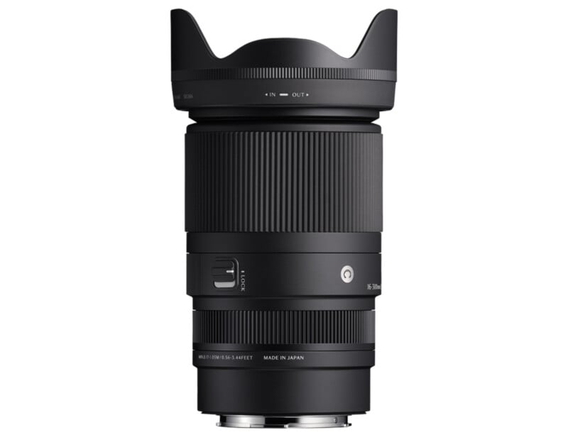 A black camera lens with a wide-angle hood is shown against a white background. It features various adjustment rings and has markings for focus and zoom settings. The lens has a polished metal mount at the bottom.