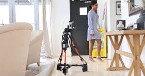 A person in a blue dress stands in a room with a camera on a tripod in the foreground. The room has a beige sofa, wooden table, and artwork on the walls.