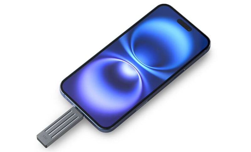 A smartphone with a blue abstract wallpaper on its screen is placed on a white surface. The phone has a sleek metallic body, and a metallic USB device is partially inserted into its port on the side.