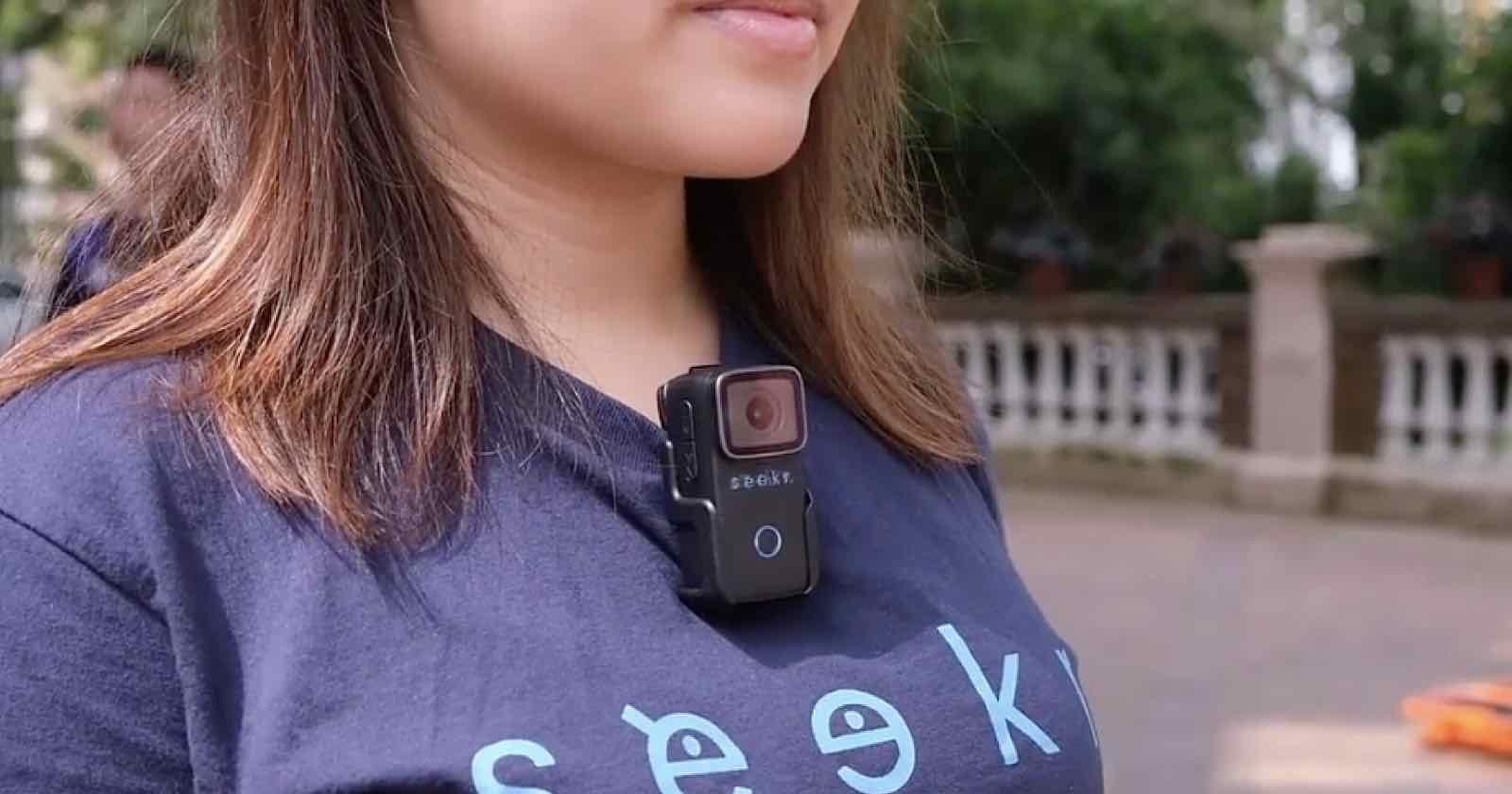 A person wearing a navy blue T-shirt with the word "seek" printed on it is standing outside. A small black camera is clipped to the front of the T-shirt. Trees and a fence are visible in the background.