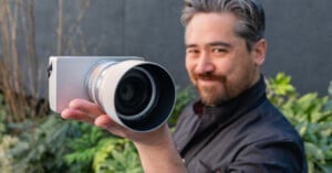 A person with gray and black hair and a beard is holding a large camera with a white lens, smiling at the camera. They are outdoors with green foliage in the background.