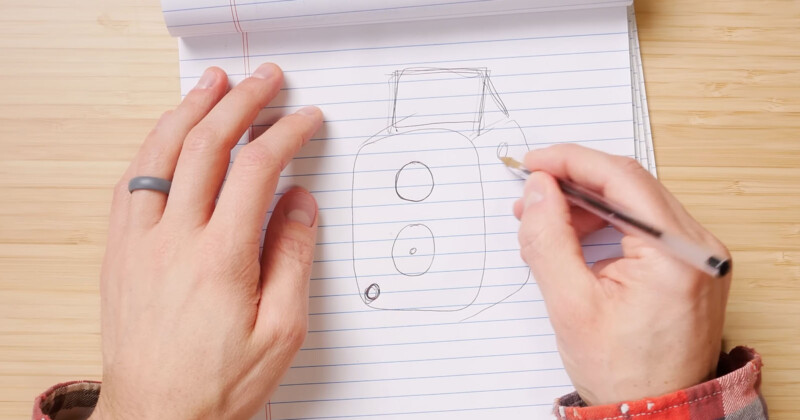 A person sketching a speaker on a lined notebook, with hands visible holding a pen and resting on the page. The notebook is open on a wooden surface, and the person is wearing a ring and a long-sleeve shirt.