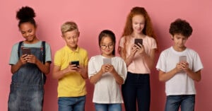 Five children standing against a pink background, each looking at their smartphone. They are dressed casually; one wears overalls, another a yellow polo, a third with glasses, wearing a peach shirt, and two others in white and beige shirts.