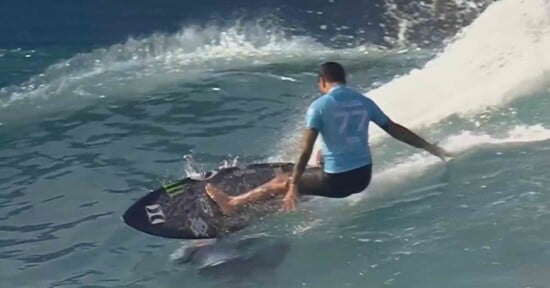 A surfer in a light blue shirt, numbered 7, rides a wave on a black surfboard. Water splashes around as they skillfully maneuver along the wave's curve. The ocean background shows clear blue water.