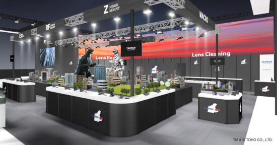 Exhibition booth featuring models of Godzilla and a cityscape, displaying lens cleaning products. Overhead signage shows "Zoom," "Macro," and other lens types. Bright screens in the background add a modern and dynamic atmosphere.