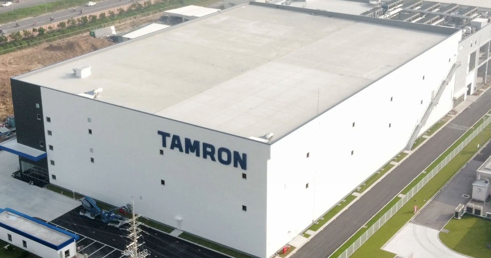 Aerial view of a large, white industrial building with "TAMRON" written in bold blue letters on the side. The building is surrounded by roads and greenery, featuring a paved area with vehicles nearby.