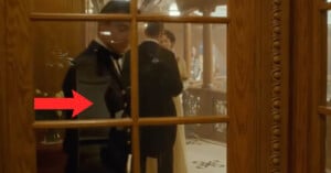 A man in a black suit and white bow tie is visible through a wooden door with glass panes. He is entering or exiting a room. A red arrow points at him. In the background, another man and a woman in formal attire are talking.