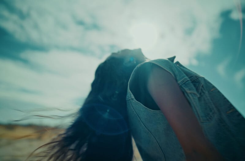 Person with long hair and sleeveless denim jacket leans back, gazing towards the bright sky. Sunlight and lens flare create a dramatic, dreamy effect, with clouds diffusing the light in a soft, blurred background.