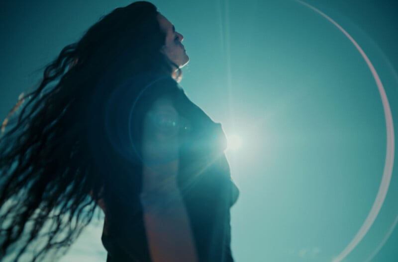 Person with long hair stands with arms outstretched toward the sky, backlit by the sun. A lens flare creates circular patterns in the blue sky, conveying a sense of freedom and serenity.