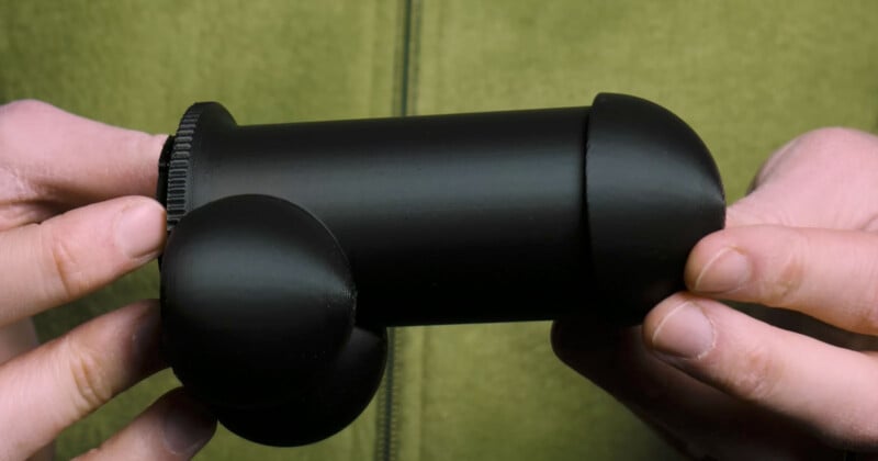 Two hands holding a black cylindrical object with rounded ends and an attached circular base on one side, against a green background. The object resembles a geometrical assembly of spheres and cylinders.