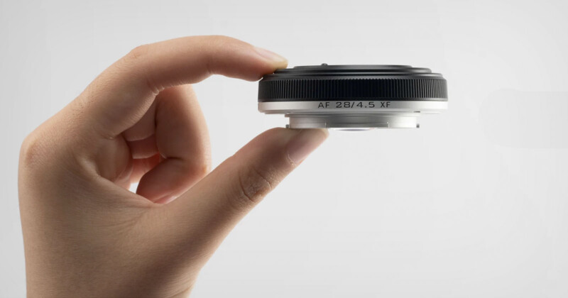 A hand holding a compact camera lens, showing a side view. The lens has a silver ring and is labeled "AF 28/4.5 XF." The background is a plain, light gray, emphasizing the sleek, minimalist design of the lens.